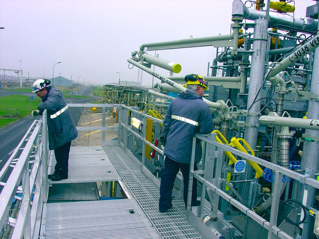 Two workers stand on the top of an elevated platform used to fuel tanker trucks.