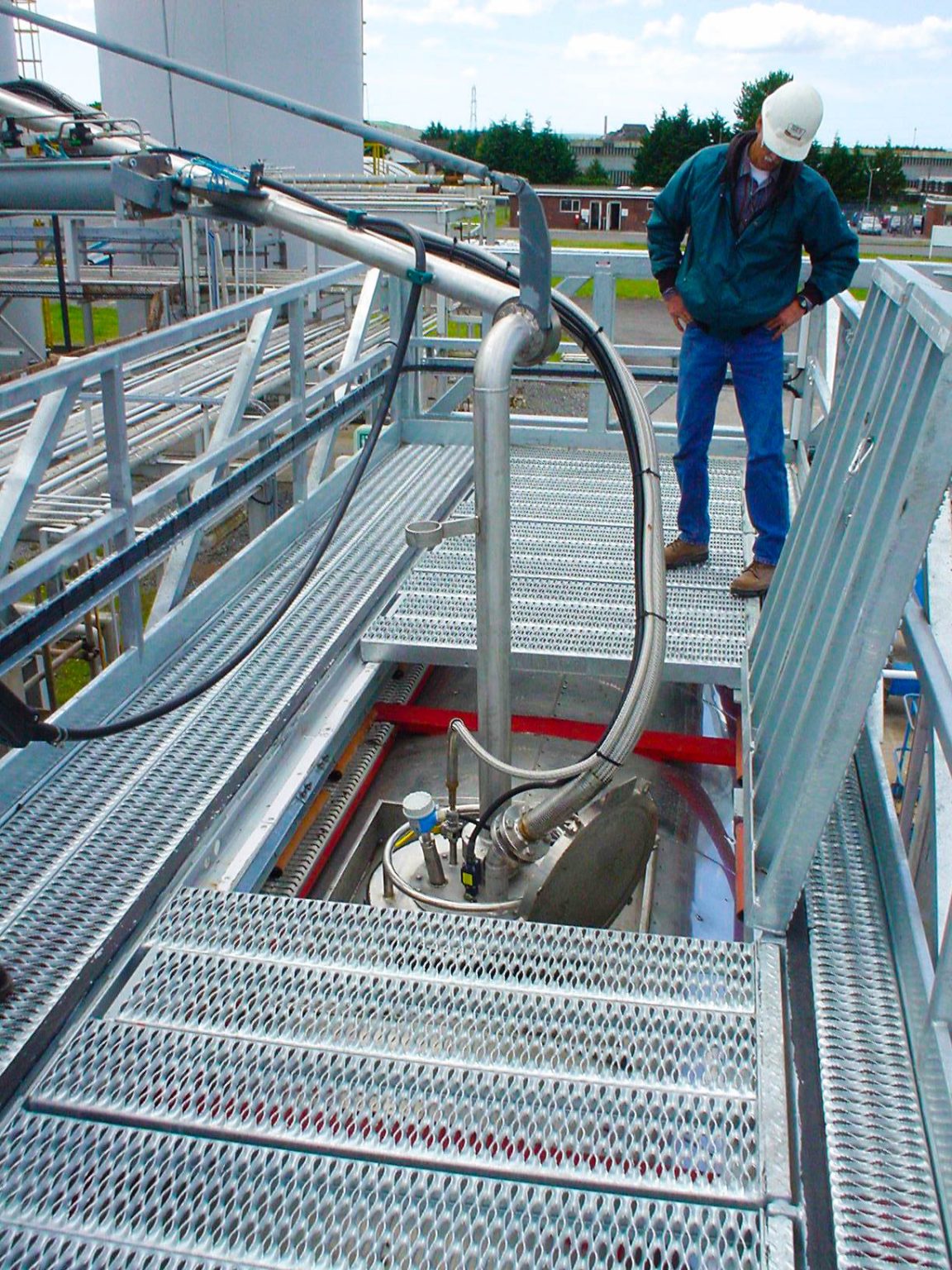 A top loading arm is shown with a safety cage over the top of a tanker railcar with a worker observing the fueling.