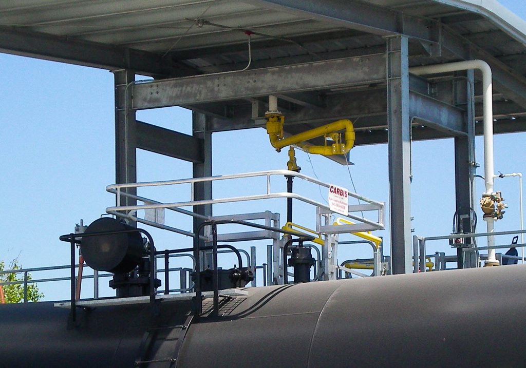 A top loading arm is shown with a safety cage over the top of a tanker railcar in a loading bay.