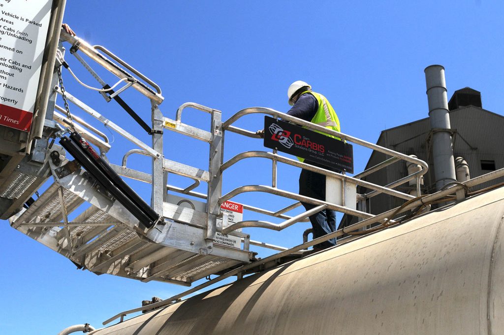 A worker stands on a Carbis Solutions folding stairs gangway enclosure above a tanker car.