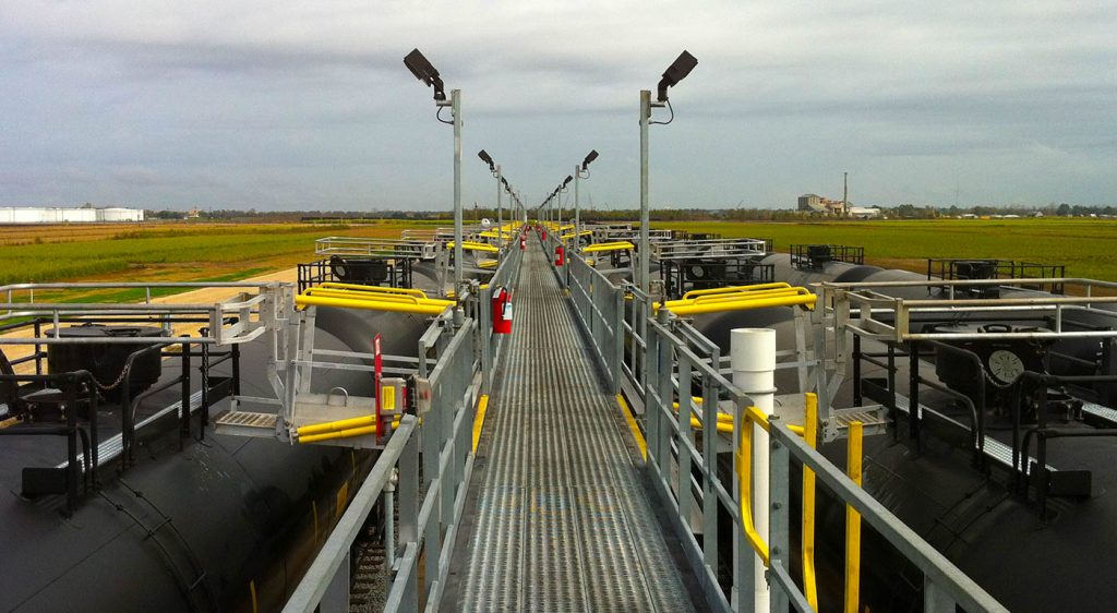 Gangways fall out on both sides of a catwalk resting above 2 railroad tracks. Railcars are accessible and underneath them.