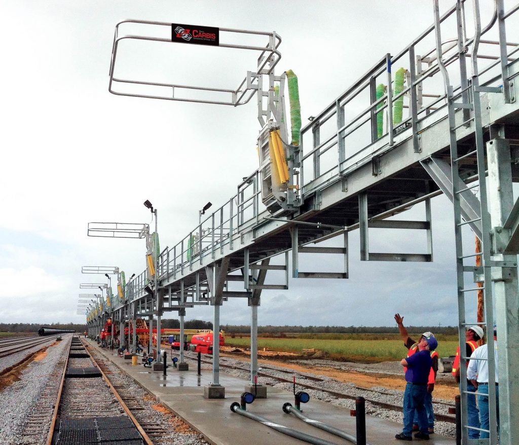 A series of railcar safety cages seen on a catwalk at a loading platform. Workers stand under them.