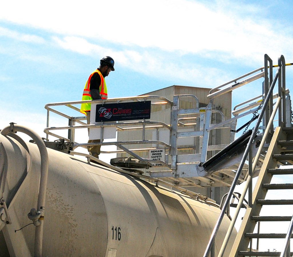 A worker stands on a Carbis Solutions folding stairs gangway enclosure above a tanker car.